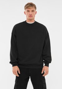 Svart crew neck sweatshirt med långa ärmar, tillverkad av mjukt tyg. Har en avslappnad passform och ribbade muddar och nederkant. Bärs tillsammans med mörka cargobyxor.