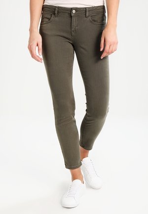 Jean slim vert olive porté par une personne debout, associé à des baskets blanches basses et un haut de couleur claire.