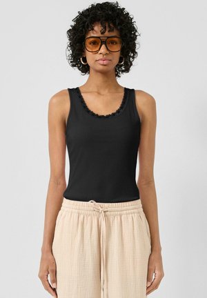 Femme aux cheveux bouclés portant de grandes lunettes de soleil ambrées, un haut noir sans manches avec bordure en dentelle et un pantalon beige clair à cordon de serrage.