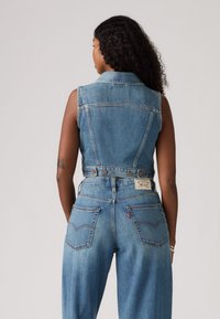 Gilet en jean avec une coupe ajustée, couleur bleu clair, fermeture à boutons, et deux poches arrières, associé à un bas en jean taille haute et à jambes larges.