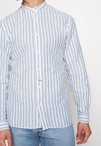 Homme portant une chemise à boutons à rayures verticales bleu clair et blanc avec col mandarin et un jean bleu clair sur fond blanc.