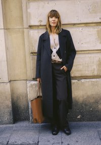 Femme portant un long manteau sombre, un cardigan à motifs, un pantalon noir, tenant un sac cabas marron, debout contre un mur en pierre beige.