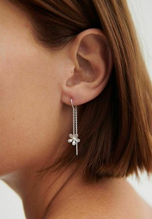 Nainen, jolla on lyhyet ruskeat hiukset ja hopeinen ketjukorvakoru, jossa on kukkakuvioinen pidike kirkkailla kivillä.