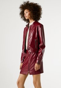 Bordeaux nep leren jasje met een kraag, ritssluiting en zijzakken, gecombineerd met een bijpassende minirok. Gladde, glanzende afwerking.