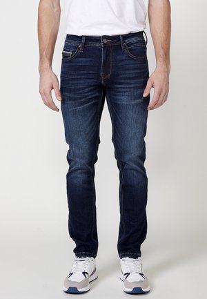 Jeans de mezclilla de color azul oscuro con un corte ajustado, que presentan un ligero efecto deslavado, cinco bolsillos y costuras en contraste. Se llevan con zapatillas blancas.