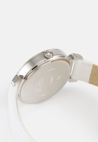 Montre en boîtier métallique argenté avec un bracelet en cuir blanc. Possède une couronne, une finition lisse et un dos uni avec des détails gravés.