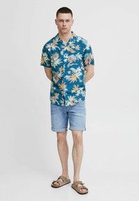 Chemisette teal à manches courtes avec un motif floral, associée à un short en denim clair et des sandales marron avec un design à boucle.