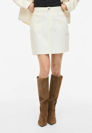 Naine, kes kannab valget teksamini seelikut ja pruune seemisnahast põlvik-saapaid valgel taustal.