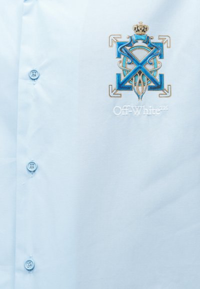 Μπροστινό μέρος ανοιχτό μπλε πουκάμισου με τρία κουμπιά και κεντημένο μπλε και χρυσό έμβλημα με κορώνα και το κείμενο "Off-White".