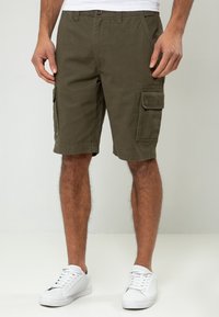 Shorts cargo vert olive en coton, dotés de poches latérales et d'une coupe régulière, associés à des baskets blanches sur un fond neutre.