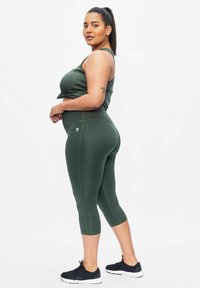 Olivgrünes Sportoberteil mit einem Knoten-Detail, kombiniert mit passenden Capri-Leggings. Verfügt über eine glatte Textur und einen Logo-Akzent am Bein.