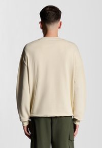 Felpa oversized color crema realizzata in morbido tessuto, con spalle cadenti e polsini a coste, indossata con pantaloni cargo verde scuro.