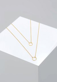 Guldfarvede halskæder med fine kædeled, der har cirkulære vedhæng. Præsenteret på en hvid geometrisk overflade mod en grå baggrund.
