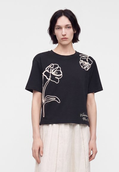 Jeune personne aux cheveux courts et foncés portant un t-shirt noir avec des motifs floraux légers en ligne et une jupe beige clair.