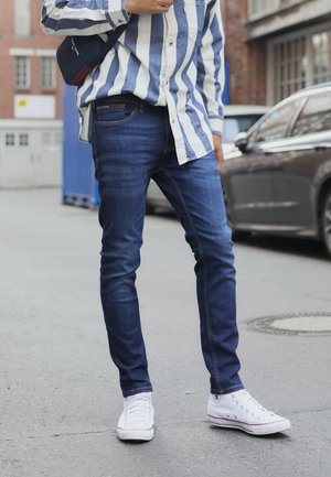Personne portant un jean skinny bleu, des baskets blanches montantes et une chemise rayée bleu et blanc à manches longues, debout dans la rue.