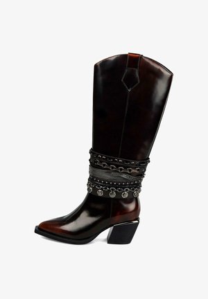 Bruine leren cowboylaars met spitse neus, versierd met decoratieve zwarte kettingen en zilveren accenten bij de enkel, met een stevige hak.