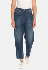 Donkerblauwe denim jeans met een losse pasvorm, voorzien van een unieke schuin afgewerkte gulp, opgerolde boorden en gecombineerd met witte sneakers.