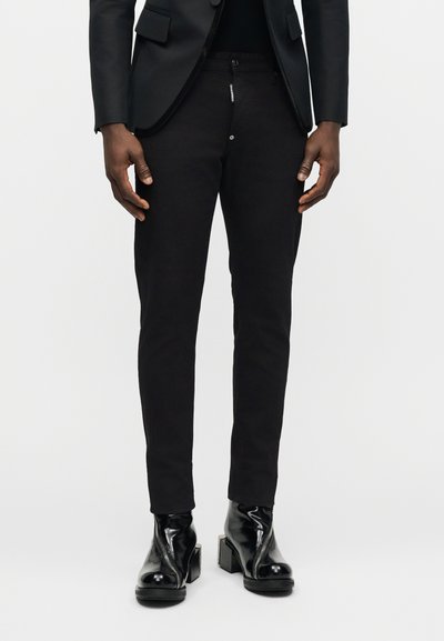 Άνδρας που φοράει στενά μαύρα παντελόνια, μαύρο σακάκι και γυαλιστερά μαύρα μποτάκια με τετράγωνες ψηλές φτέρνες, στέκεται με τα χέρια χαλαρά στα πλάγια.