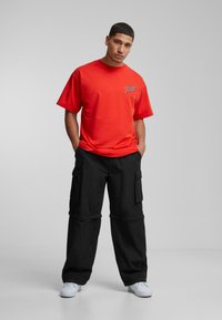 Rød bomulds t-shirt med et trykt grafisk motiv på brystet; parret med løstsiddende sorte cargo bukser med sidelommer og hvide sneakers.