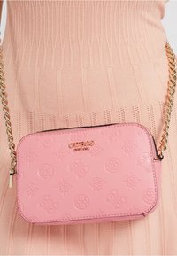 Lys rosa crossover-taske med præget mønster og guld GUESS-logo, båret over en ferskenfarvet ribstrikket kjole med geometrisk tekstur.