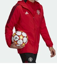 Chaqueta roja con cremallera y detalles en negro, que presenta el logo del Manchester United; sosteniendo un balón de fútbol con una base blanca y un patrón de estrellas rojas.