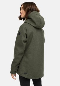 Femme avec des cheveux bruns arrivant aux épaules, portant un manteau d'hiver à capuche vert olive et un pantalon noir, se tenant de profil contre un fond uni.