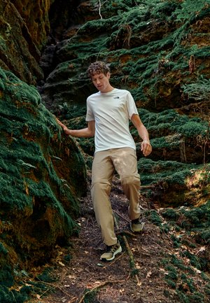 Conjunto informal que incluye una camiseta blanca de manga corta con un logo, pantalones beige y botas de senderismo oscuras. Rodeado de un terreno verde y musgoso.