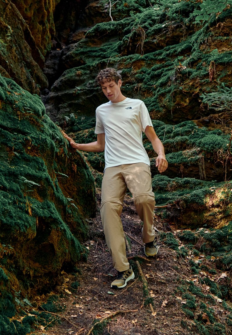Conjunto informal que incluye una camiseta blanca de manga corta con un logo, pantalones beige y botas de senderismo oscuras. Rodeado de un terreno verde y musgoso.