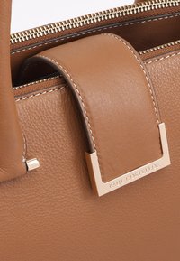 Bruine leren handtas met een zachte textuur, voorzien van een opvallende vierkante hardware-accent en contrasterende witte stiksels.