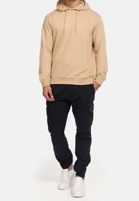 Sweat à capuche beige avec cordons de serrage, associé à un pantalon cargo noir avec poches latérales et poignets élastiques. Des baskets blanches complètent la tenue.