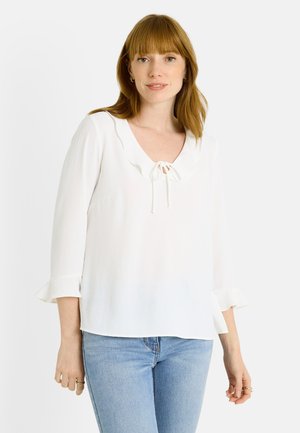 Blusa blanca con cuello festoneado y detalle de lazo, con mangas de tres cuartos y acentos de volantes. Tejido suave y ligero.