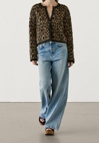 Kardigan med leopardmönster och svarta knappar, i kombination med ljusblå vida jeans. Modellen bär platåsandaler. Bakgrunden är enfärgad och neutral.