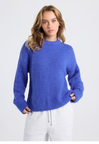 Blauer, flauschiger Strickpullover mit Rundhalsausschnitt, lockerem Schnitt und gerippten Bündchen. Wird über grauen Jogginghosen getragen und zeichnet sich durch eine strukturierte Stoffqualität aus.