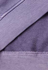 Capas de tela de color púrpura claro: una de punto suave con un dobladillo acanalado, la otra texturizada. Costuras visibles que destacan y una apariencia suave.