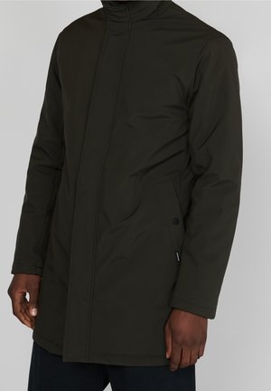 Mężczyzna ubrany w ciemnozieloną, zimową kurtkę z długim rękawem, wysokim kołnierzem i bocznymi kieszeniami na jednolitym tle.