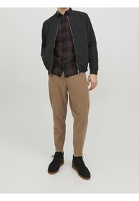 Giacca bomber nera con zipper, camicia a quadri in tonalità marroni, pantaloni in corduroy beige e scarpe in suede nere con suola marrone.