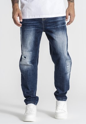 Mand iført mørkeblå slidte jeans og hvide sneakers, stående mod en ensfarvet lys baggrund, hænder afslappet langs siden.