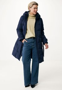 Cappotto piumino blu navy con cappuccio, caratterizzato da una vita cintrata, abbinato a un maglione a trecce color crema e jeans a gamba larga blu. Texture del tessuto liscia.