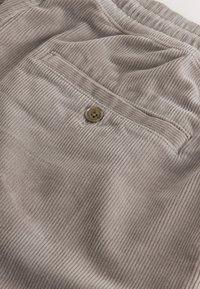 Gros plan sur un tissu en velours côtelé beige montrant une poche avec un seul bouton marron et des détails de couture visibles.