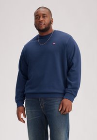 Navy cotton sweatshirt with ribbed cuffs and hem, featuring a small red logo patch. Paired with blue denim jeans.