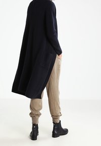 Larga chaqueta de punto azul marino con textura acanalada, bolsillos traseros y aberturas laterales. Combinada con pantalones beige y botines negros.