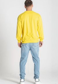 Sudadera amarilla de manga larga con puños y dobladillo acanalados, combinada con jeans de color azul claro, llevada con zapatillas blancas. Fondo liso.