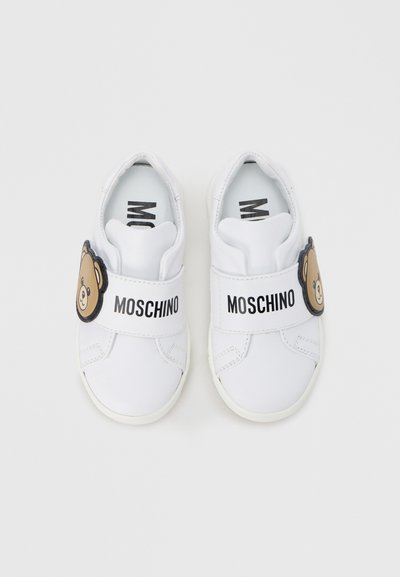 Λευκά πάνινα παπούτσια τύπου slip-on με λαστιχένια σόλα, που διαθέτουν μαύρο λουράκι και σχέδιο αρκούδας. Το όνομα της μάρκας «Moschino» τυπωμένο στο λουράκι.