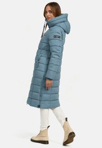 Abrigo acolchado hasta la rodilla en azul claro con capucha, cierre de cremallera, bolsillos frontales y un diseño acolchado. Combinado con botas beige con cordones.