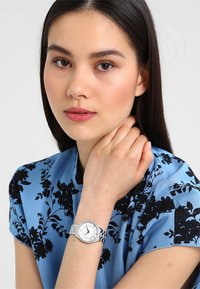 Silberne Uhr mit einem runden weißen Zifferblatt und schwarzen Ziffern, am Handgelenk getragen. Das Display besitzt ein Edelstahlarmband und eine gemusterte blaue Bluse mit Blumenmuster.
