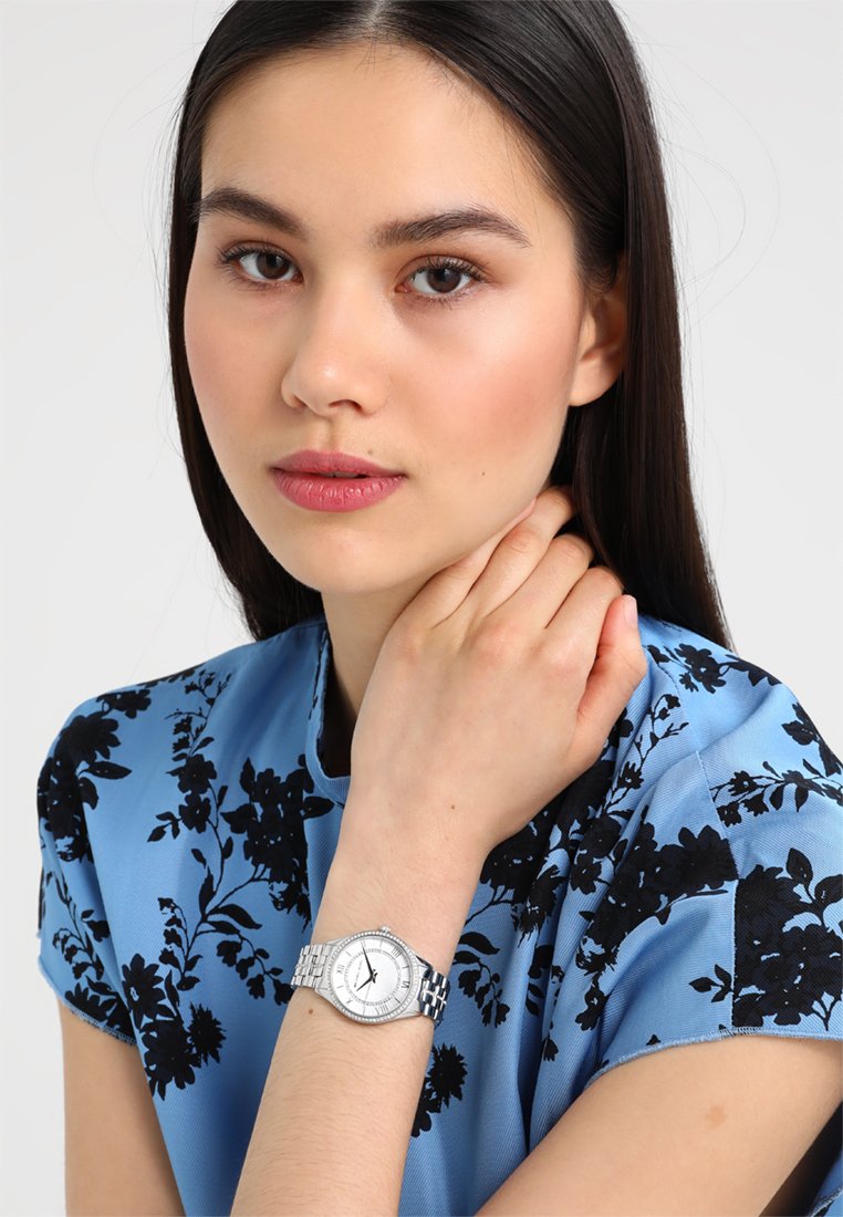 Silberne Uhr mit einem runden weißen Zifferblatt und schwarzen Ziffern, am Handgelenk getragen. Das Display besitzt ein Edelstahlarmband und eine gemusterte blaue Bluse mit Blumenmuster.