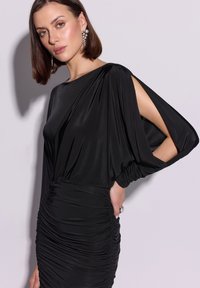 Czarna sukienka z lekkiego materiału, z długimi rękawami w stylu drapowanym, dopasowanym gorsetem i marszczeniem w talii.