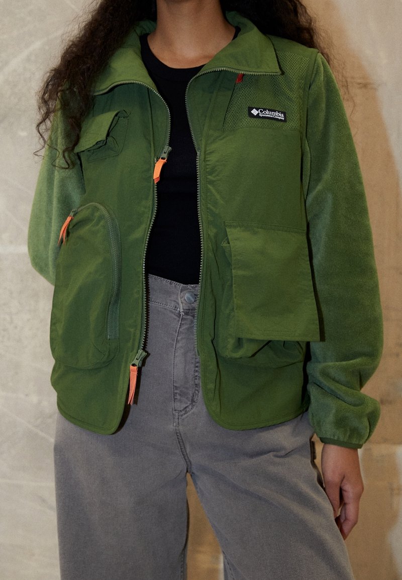 Zelená fleecová bunda se zipem v přední části, několika kapsami a oranžovými jezdci na zip. Má vysoký límec a žebrované manžety, nošena přes šedé kalhoty.