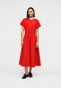Κόκκινο βαμβακερό φόρεμα με στρογγυλή λαιμόκοψη, κοντά μανίκια και ζωνάρι στο ύψος της μέσης που ανοίγει σε φουσκωτή φούστα. Μαύρα loafers συμπληρώνουν την εμφάνιση.