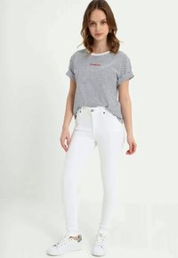 Jovem mulher vestindo uma t-shirt cinza e branca com riscas, jeans skinny brancos de cintura alta e tênis brancos, em pé contra um fundo neutro.